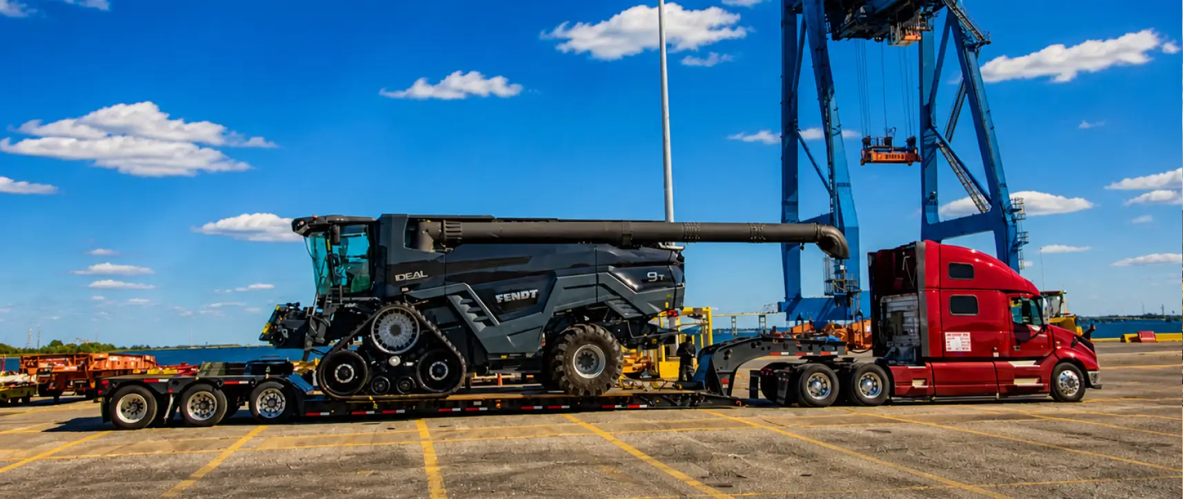 Oversize combine harvester transported on a flatbed trailer by a heavy haul truck at a port with crane in background
