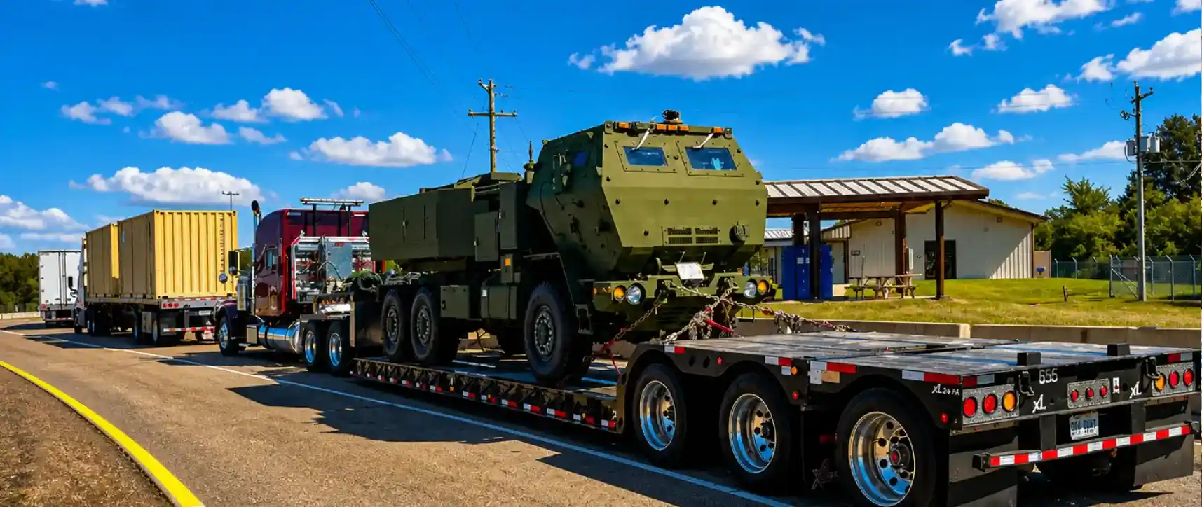 Vehículo militar sobredimensionado transportado en un remolque de plataforma por un camión de carga pesada en carretera bajo cielo azul despejado